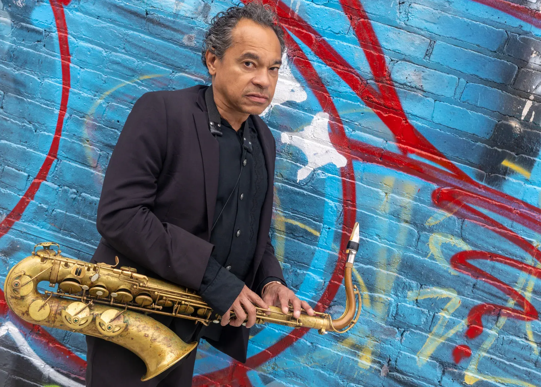 Jay Rodriguez Sierra with saxophone in front of a graffiti wall.
